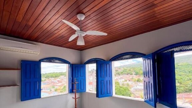 A room with wooden ceiling, a ceiling fan, two beds with white and purple bedding, and blue shutters on the windows. - Pousada Lavramor (Lençóis)