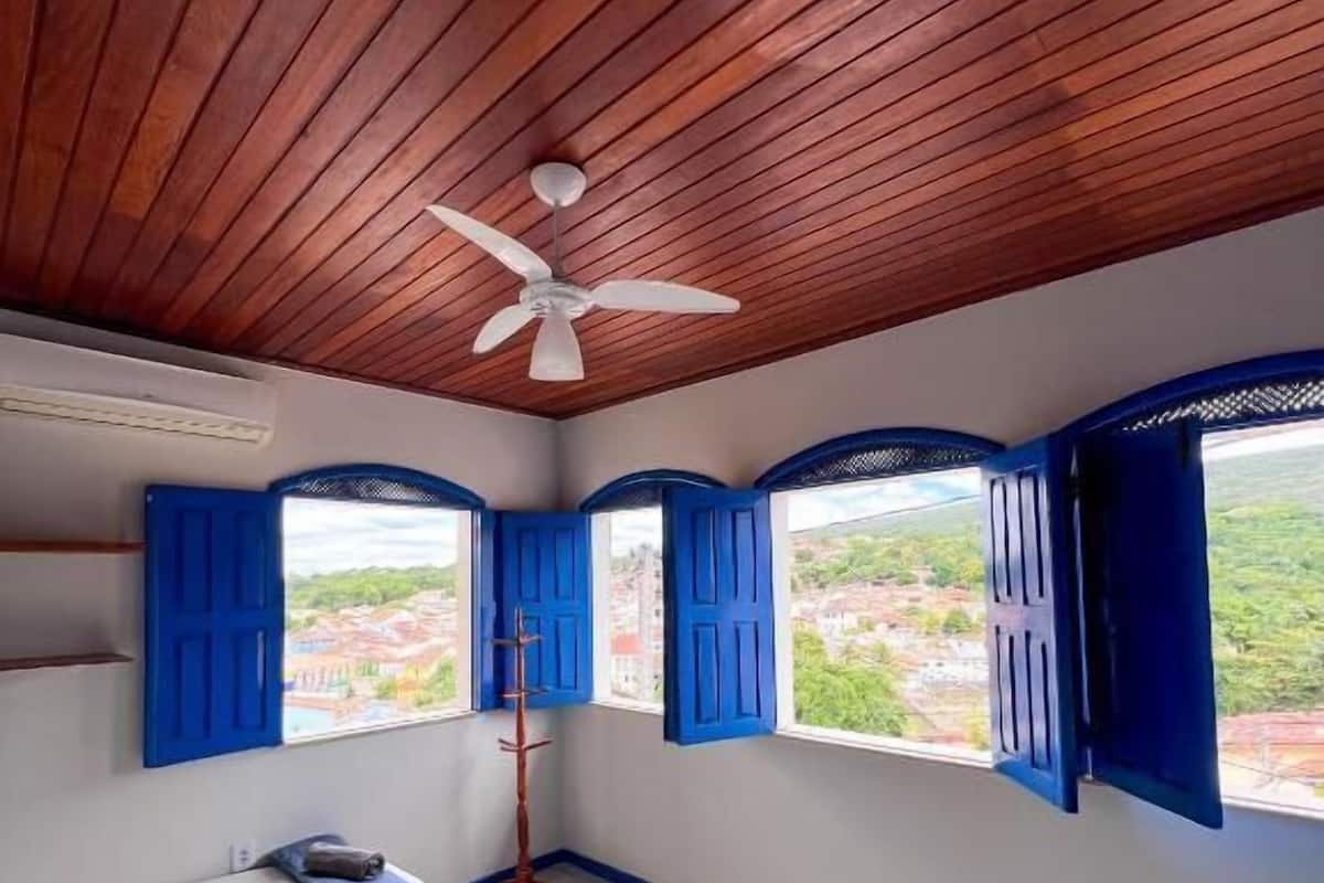 A room with wooden ceiling, a ceiling fan, two beds with white and purple bedding, and blue shutters on the windows.