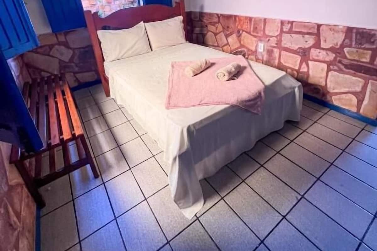 A single bed with a pink pillow and a wooden bench in a room with stone accents on the lower wall and blue curtains.