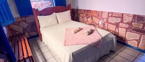 A single bed with a pink pillow and a wooden bench in a room with stone accents on the lower wall and blue curtains.