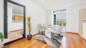 Un espace de repas moderne avec une table et des chaises en verre, un grand miroir, une plante en pot et une vue sur le paysage urbain à travers de grandes fenêtres.
