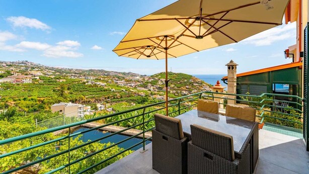 Une terrasse avec une table et des chaises, protégée par un grand parasol, d’où l’on peut admirer un vignoble et une ville côtière.