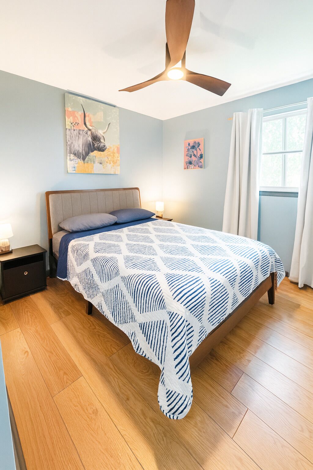Un dormitorio con cama, mesita de noche, ventilador de techo y obras de arte en las paredes.