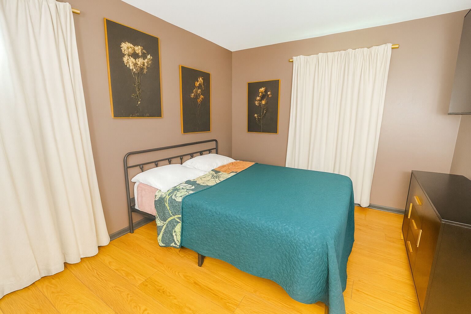Une chambre à coucher comprenant un lit, des rideaux, une commode et trois œuvres d’art florales encadrées accrochées au mur.