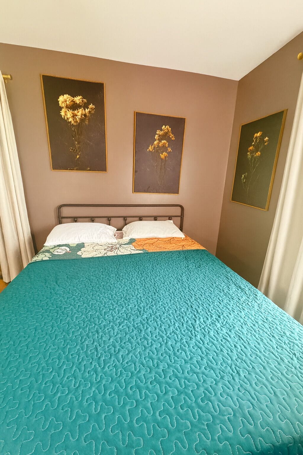 Ein Schlafzimmer mit einem Bett, einer türkisnen Steudecke und drei gerahmten floralen Bildern an der Wand.