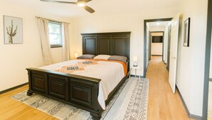 Ein Schlafzimmer mit einem dunklen Holzbett, einem Deckenventilator, einem Fenster mit Vorhängen und einem zur anderen Zimmer führenden Gang.