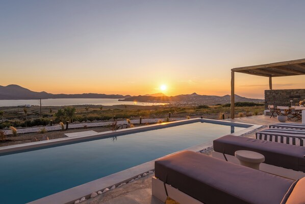 A poolside area with lounge chairs and a view of the sunset over the sea.