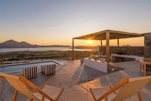 A poolside area with a view of the sunset over the water, outdoor seating, and a stone wall.