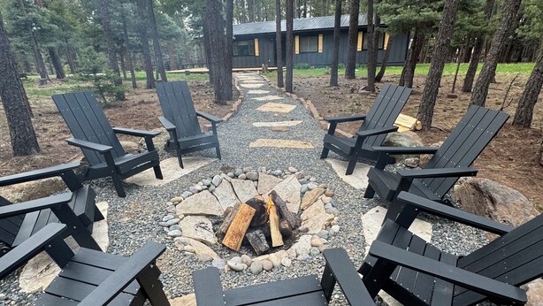 Un espace de détente circulaire, composé de fauteuils Adirondack disposés autour d’un foyer central, situé dans une zone boisée avec un bâtiment en arrière-plan.