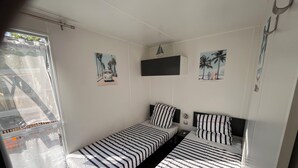 A small, well-lit room with two single beds, a window with a view of palm trees, and a wall-mounted TV. - Mobilhome cap ferret (Lège-Cap-Ferret)