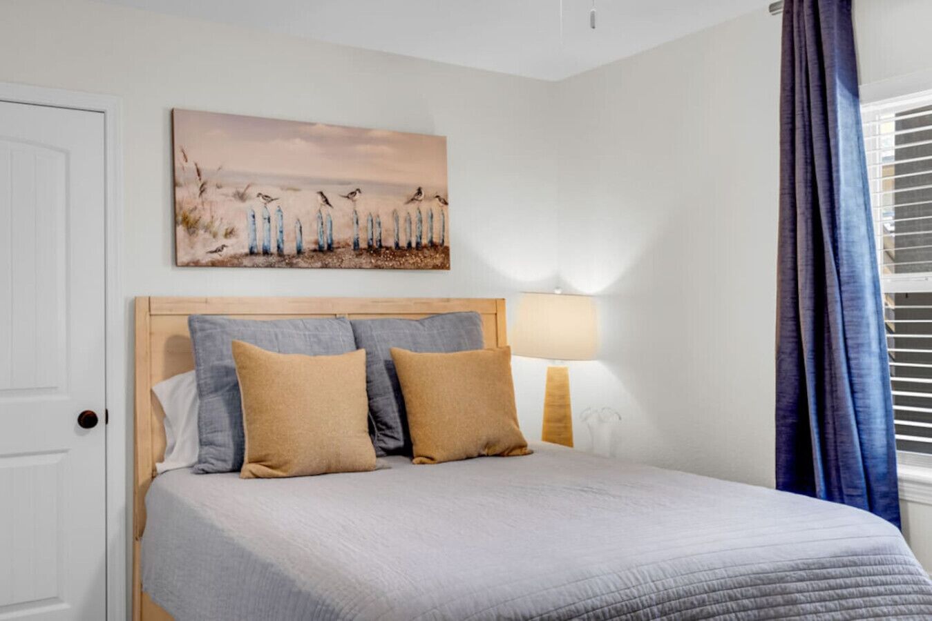 A bedroom with a bed, two pillows, a bedside lamp, a painting on the wall, and a window with curtains.