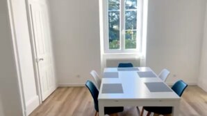Una habitación con una mesa blanca y sillas azules, una ventana con vistas a árboles, y suelo de madera.