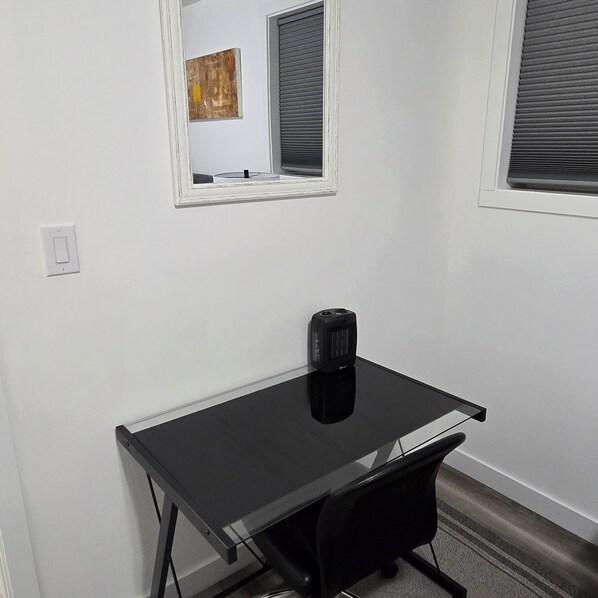 A small, modern room with a black desk, a chair, a small black heater, a white wall, and a window with blinds.
