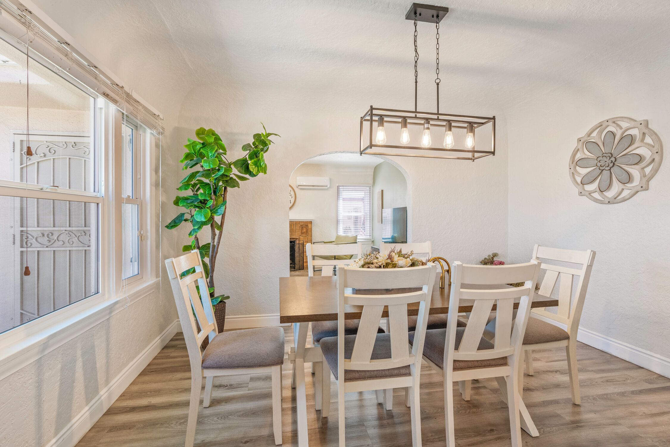 A dining area with a wooden table and white chairs, a hanging light fixture, and a large green plant.