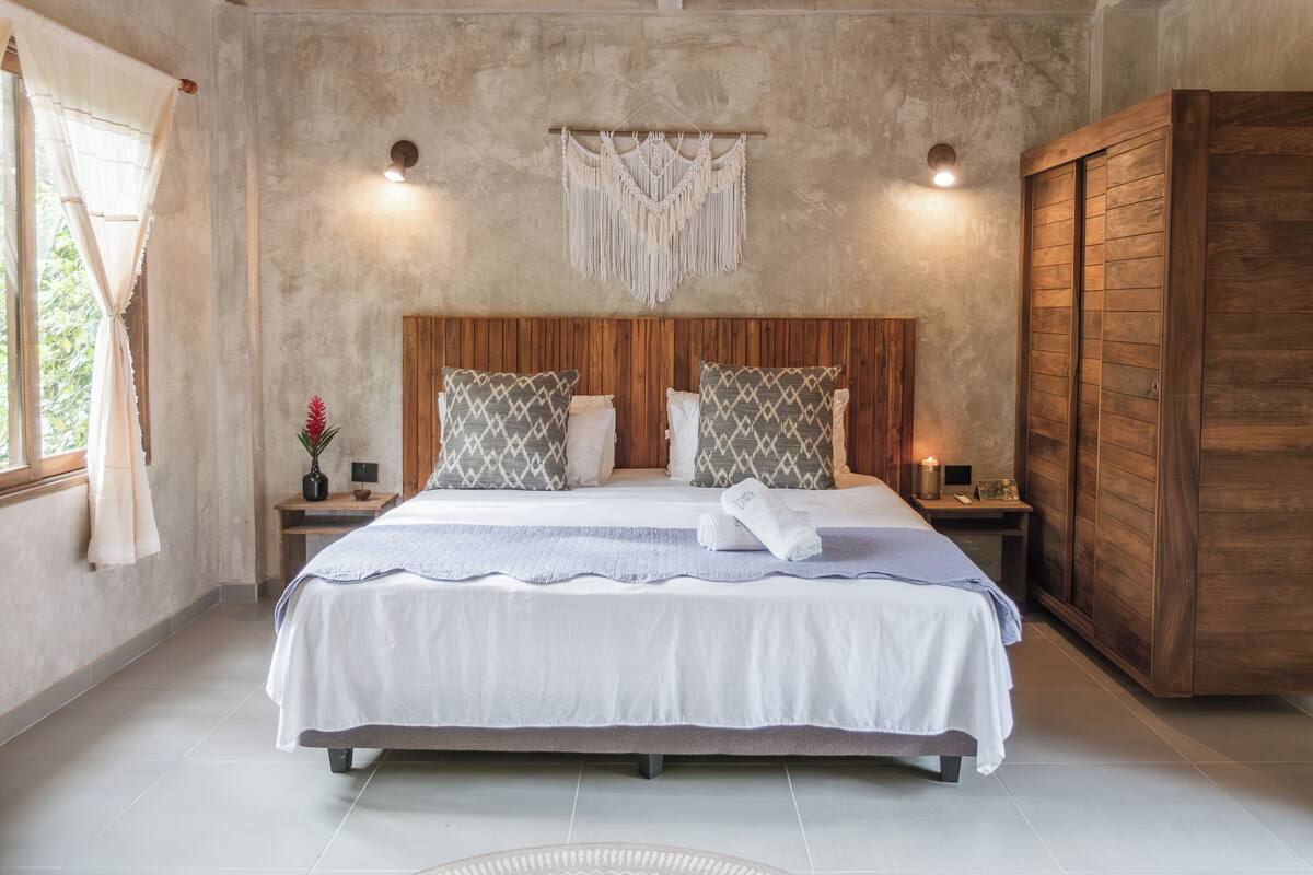 Quarto com cabeceira de madeira, cama grande com almofadas estampadas, um pendente de macramé na parede e um guarda-roupa de madeira.