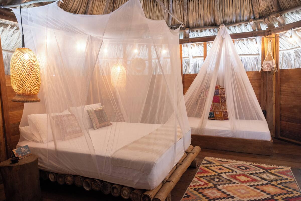 Quarto com duas camas, cada uma coberta por um dossel branco. Há uma cabeceira de madeira, um cesto de fibra natural e um tapete estampado no chão.