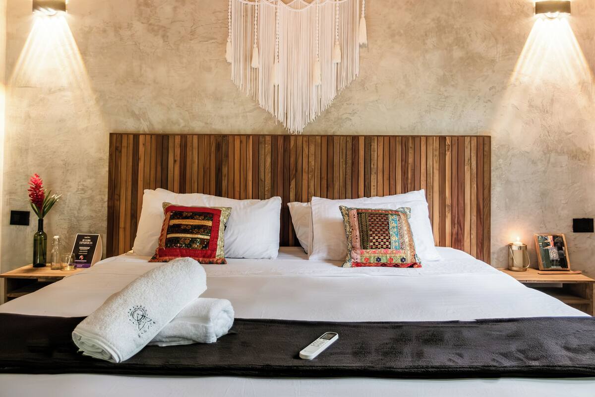 Quarto de hotel com cabeceira de madeira, roupa de cama branca, almofadas decorativas, um pendente de macramé na parede e mesas de cabeceira com luminárias e um vaso de flores.
