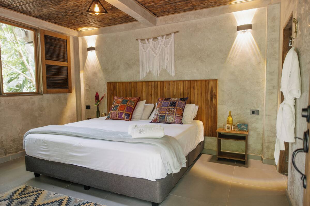 Quarto com cama grande, cabeceira de madeira e uma pequena mesa de cabeceira com uma garrafa e um livro.