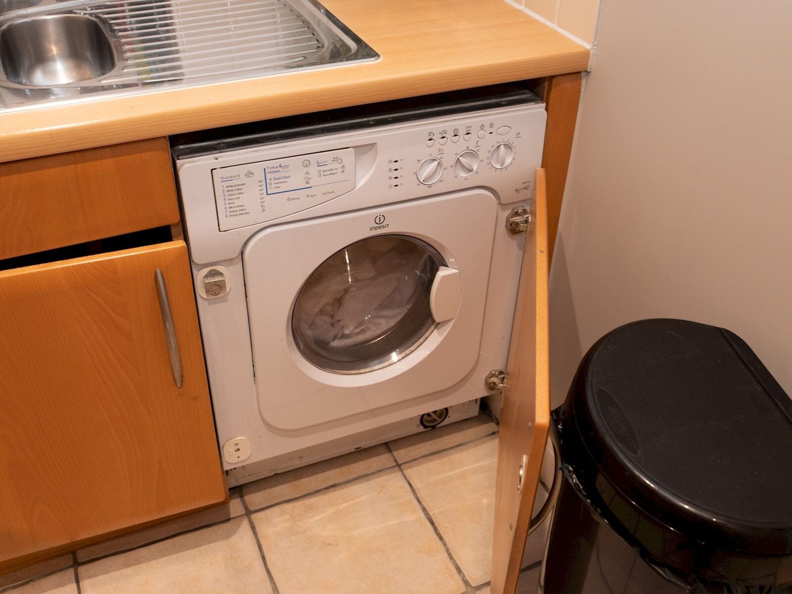 Een compacte wasmachine met bedieningspaneel, een wasbak en een prullenbak in een kleine keuken.
