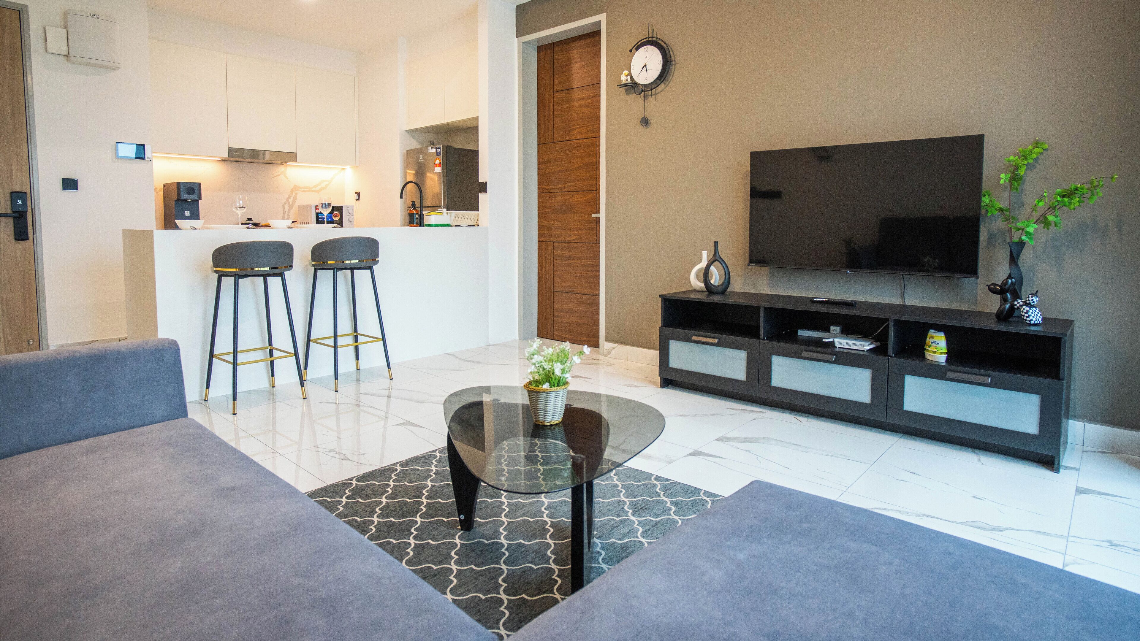 A modern living room with a glass coffee table, grey sofas, a TV, and a kitchen in the background.