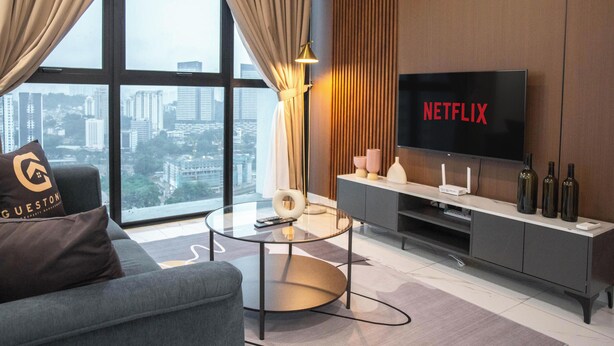 A modern living room with a grey sofa, a glass coffee table, a flat-screen TV displaying 'NETFLIX', and a city view through large windows.