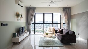 Una sala moderna con un sofá, una mesa de centro y un televisor sobre un mueble bajo. Hay un ventanal con cortinas, un ventilador de techo y vistas a un paisaje urbano.
