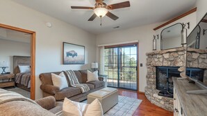 A cozy living room with a stone fireplace, a ceiling fan, and a large window with a view of the outdoors. - 2BR Village At Indian Point 1202 near SDC-Free Tickets Daily up to $500 (Branson)