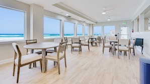 Un vaste espace dédié à la salle à manger, doté de grandes fenêtres donnant sur l’océan, de tables et de chaises en bois, ainsi que d’un ventilateur de plafond.