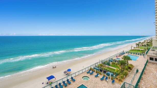 Une vue sur la plage depuis le balcon d’un hôtel, avec une piscine, des chaises longues et un ciel bleu dégagé.