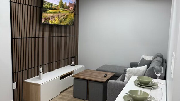 Una sala moderna con televisor de pantalla plana montado en una pared de paneles de madera, un sofá gris, una mesita de centro pequeña y una zona de comedor con una mesa blanca y cuencos verdes.