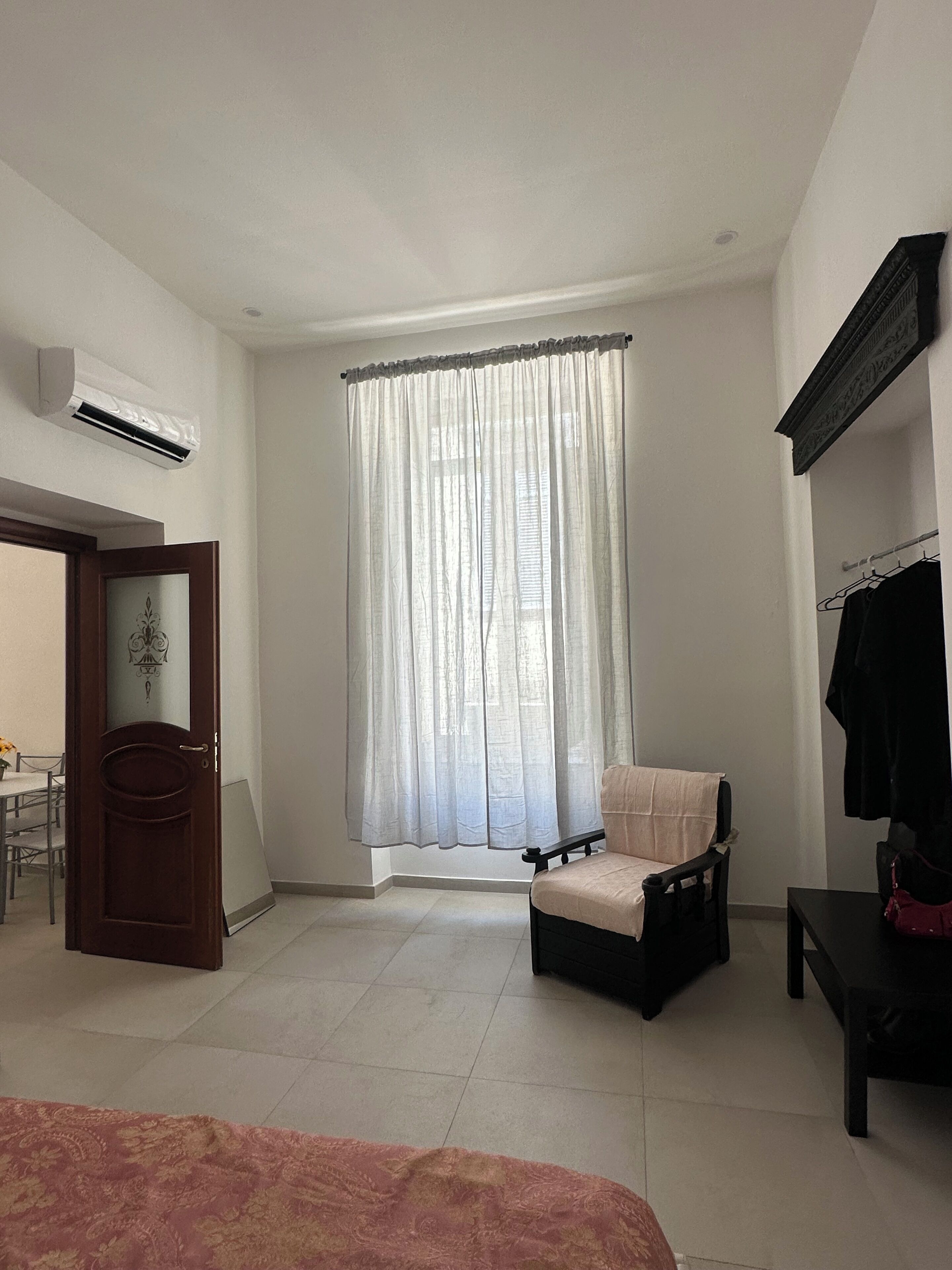 A bedroom with a bed, a chair, a small table, a coat rack, and a window with sheer curtains.