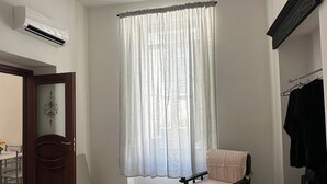 Une chambre à coucher comprenant un lit, une chaise, une petite table, un porte-manteau et une fenêtre avec des rideaux légers.