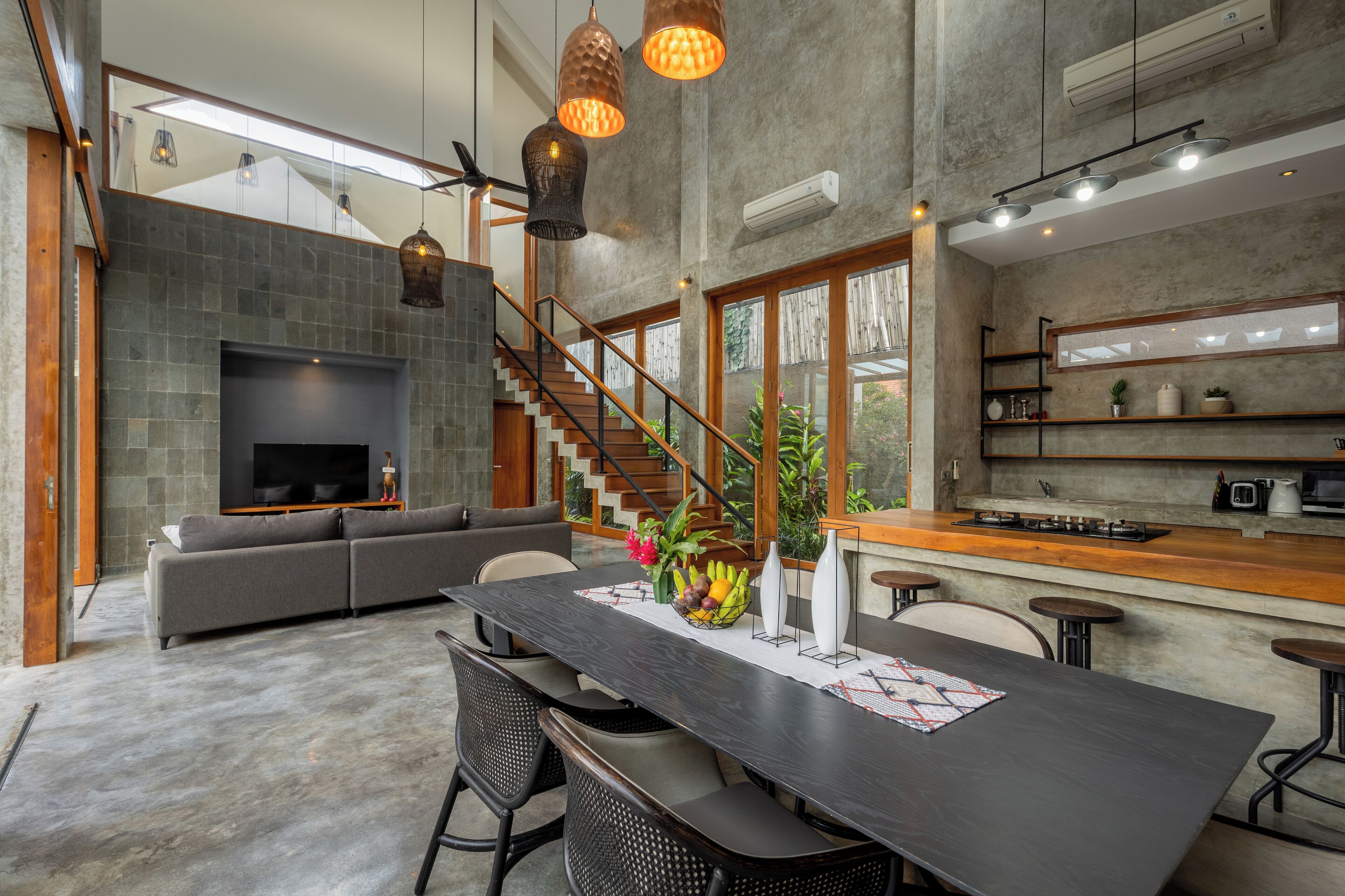Una sala de estar moderna con una mesa de comedor de madera oscura, un sofá gris y un televisor. Una escalera conduce a un nivel superior.