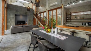 Ein modernes Wohnzimmer mit einem dunkelholznen Esstisch, einem grauen Sofa und einem Fernseher. Eine Treppe führt zu einer oberen Ebene.