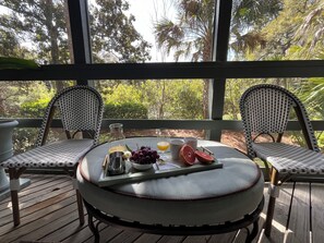 A round table with a tray, two wicker chairs, a bottle, a bowl of grapes, a grapefruit, and a glass of orange juice on a wooden deck with a view of trees.