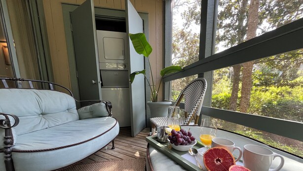 A cozy room with a curved sofa, a dining table with breakfast items, a view of trees through large windows, and a kitchen area. - One Block Kiawah Beach Villa (Kiawah Island)