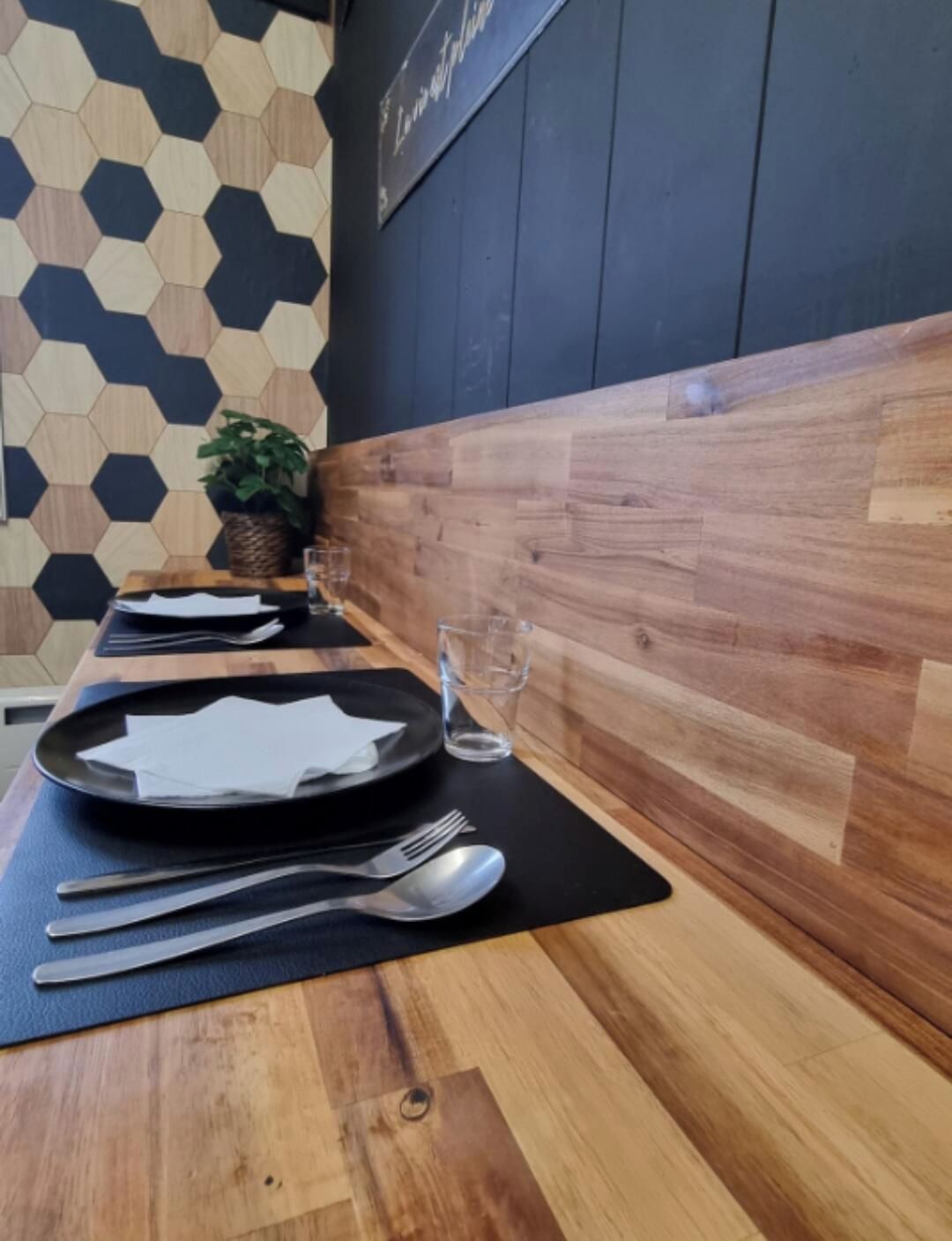 Una mesa de comedor dispuesta con platos, cubiertos y vasos, con una pared de madera y un patrón geométrico al fondo.