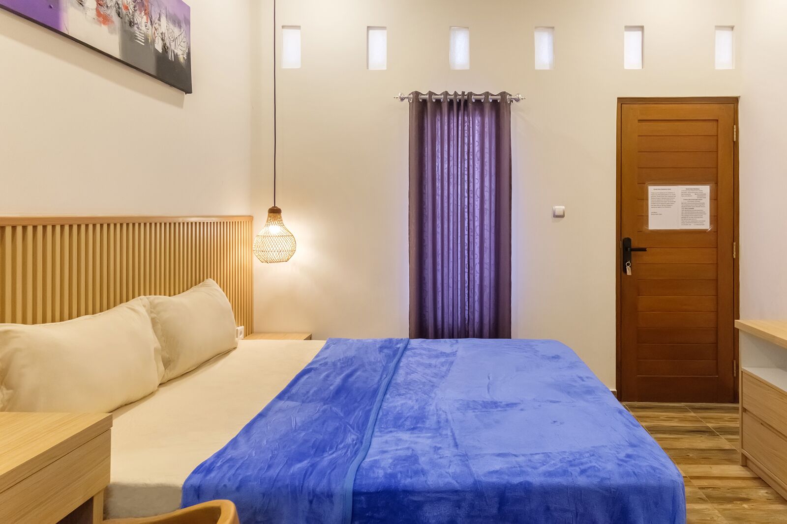 Een slaapkamer met een bed, een nachtkastje, een houten hoofdeinde, een hanglamp, een deur met een bord en een raam met gordijnen.