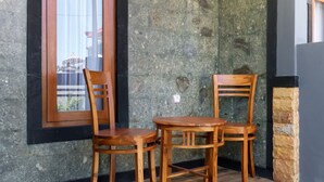Een hoekje met twee houten stoelen en een tafel, een raam met uitzicht op gebouwen, en een muren van ruw natuursteen.