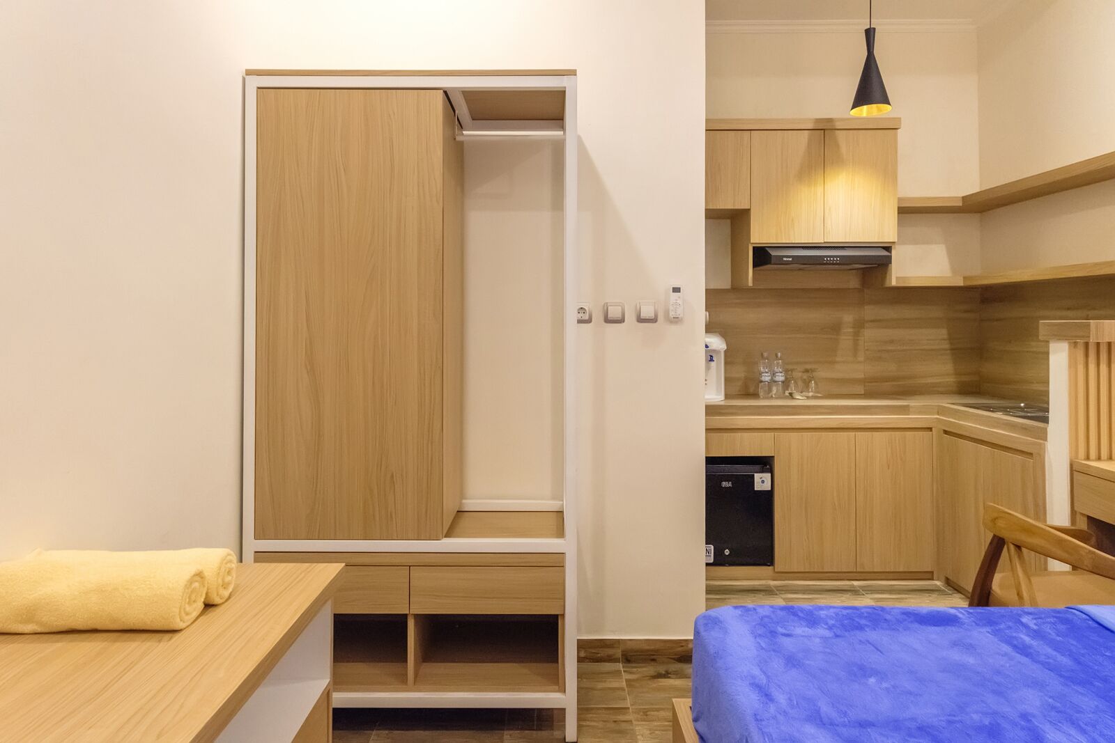Habitación compacta con cama, una mesita con una toalla, kitchenette con armarios de madera, refrigerador negro y una lámpara colgante.