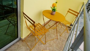 Ein gelber Tisch und zwei Holzstühle mit einem Blumentopf darauf, platziert auf einem Balkon mit Glastür und Metallgeländer.