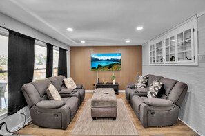 Un salon moderne avec deux canapés inclinables, un pouf central et un grand téléviseur à écran plat fixé à un mur en bois.