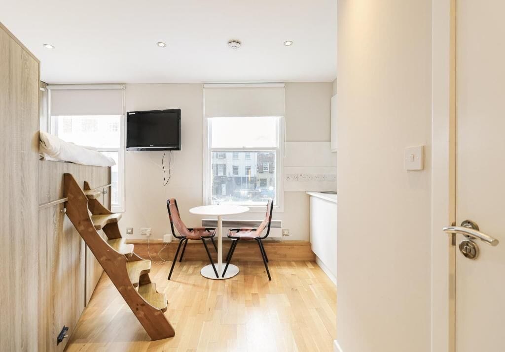 A modern interior with a staircase, a dining table with chairs, a flat-screen TV mounted on the wall, and a window with a city view.