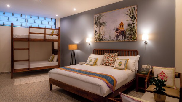 A hotel room with a large bed, a smaller bunk bed, a nightstand, a lamp, a wall painting, and a potted plant. - Inara House (Agra)