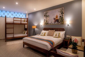 A hotel room with a large bed, a smaller bunk bed, a nightstand, a lamp, a wall painting, and a potted plant.