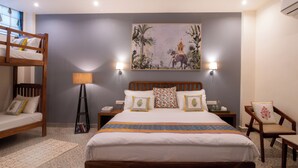 A hotel room with a bunk bed, a large bed with multiple pillows, a nightstand with a lamp, a chair, and a wall-mounted air conditioning unit. - Inara House (Agra)