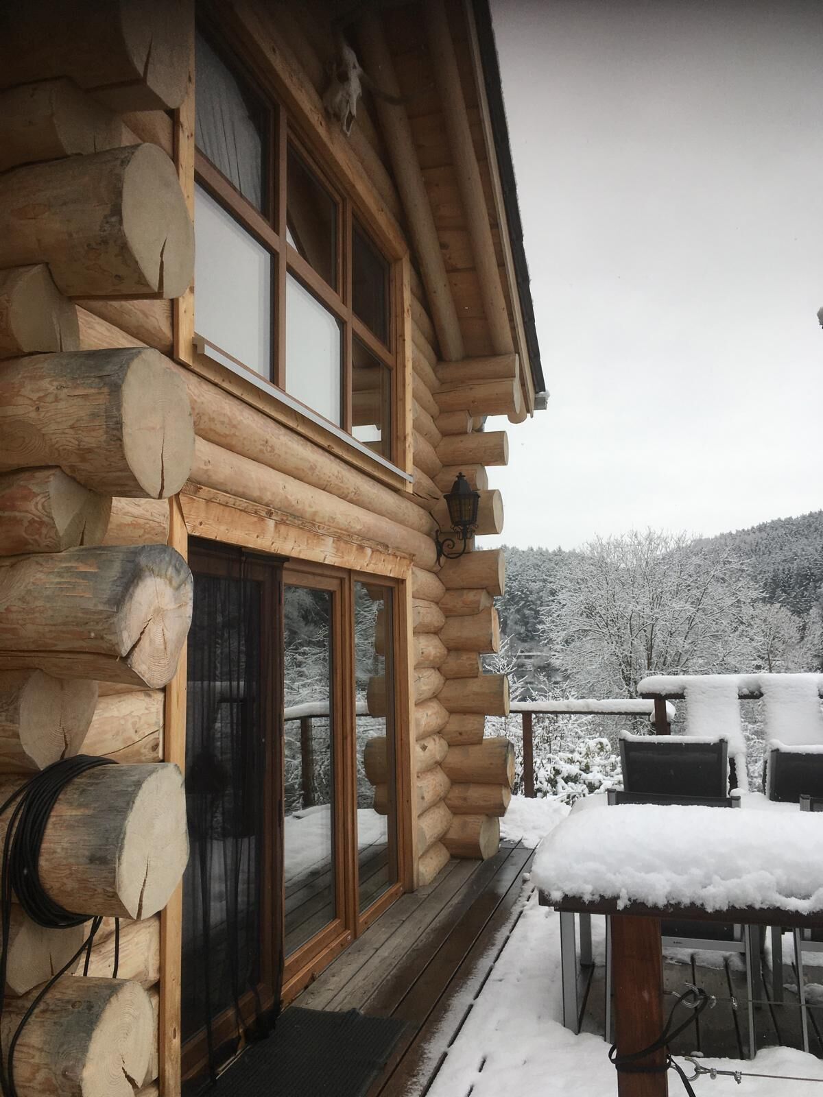 A log cabin with a wooden deck, large windows, and a view of a snowy landscape.