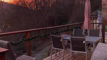 A wooden deck with outdoor furniture, including chairs and a table, overlooking a forested area with a sunset in the background.