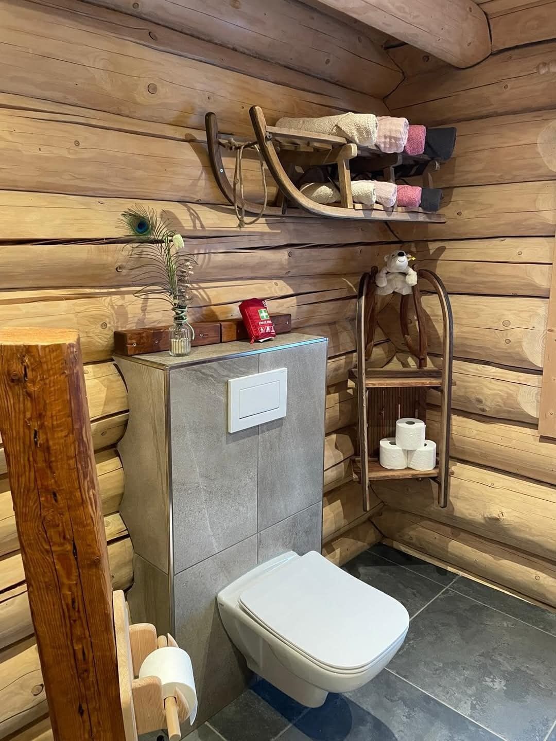 A compact bathroom with a toilet, a shelf with toiletries, and a wooden bench.