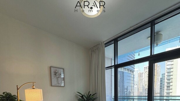 A modern living room with a sofa, armchairs, a coffee table, and a floor lamp. There is a large window with a view of buildings. - 1BR Near metro.Canal  View. Mer. 1709 (Dubai)
